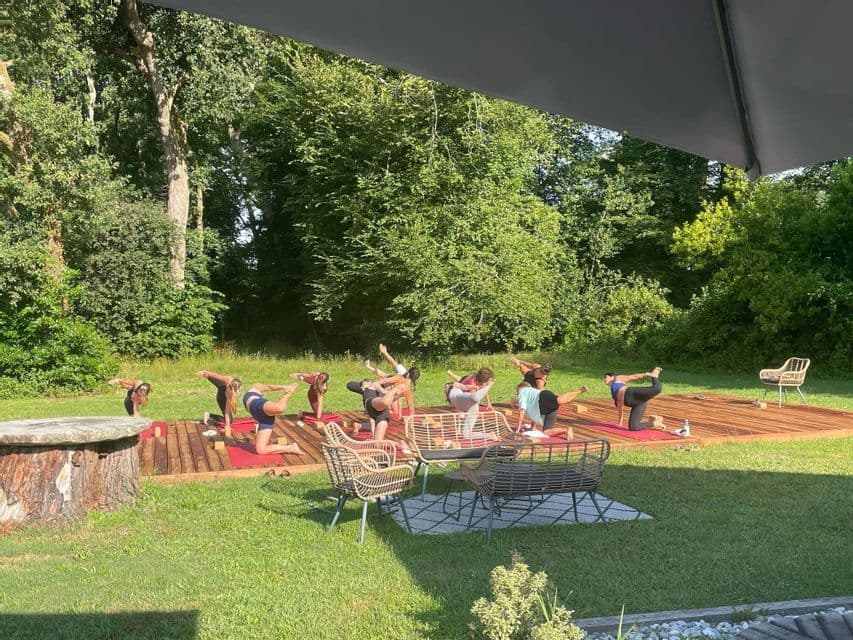Un groupe WeRoad pratique le yoga sur des tapis rouges sur une terrasse en bois dans un jardin verdoyant et arboré.