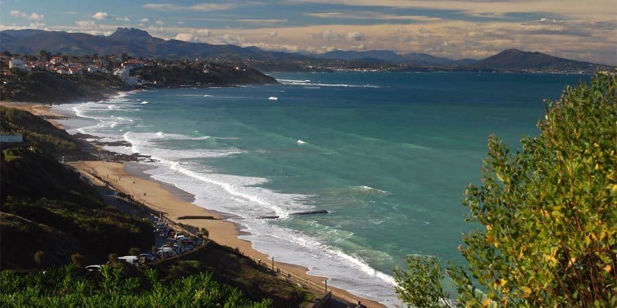 Vue surélevée d'une côte sinueuse avec une plage de sable, une eau turquoise, une ville balnéaire et des montagnes sous un ciel bleu.