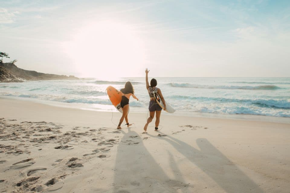 Deux personnes d'un voyage de groupe WeRoad portant des planches de surf marchent le long d'une plage de sable vers les vagues de l'océan au lever du soleil.