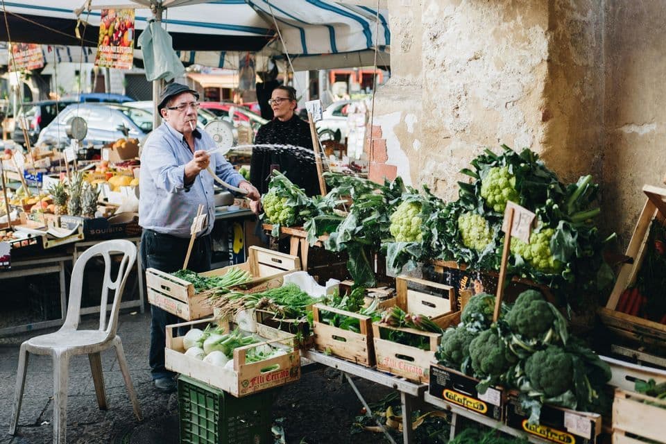 Un venditore in una bancarella del mercato all'aperto spruzza acqua con una canna su cassette di broccoli e cavolfiori freschi.