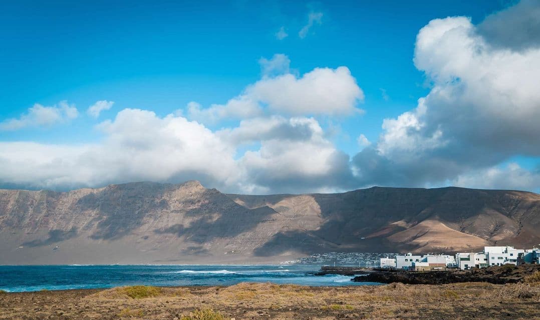 Un borgo costiero con case bianche sorge sul mare, ai piedi di grandi montagne, sotto un cielo parzialmente nuvoloso.