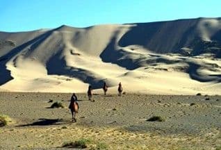 Un viaggio di gruppo WeRoad si sposta a cavallo attraverso una pianura arida, diretto verso enormi dune di sabbia sotto un cielo sereno.