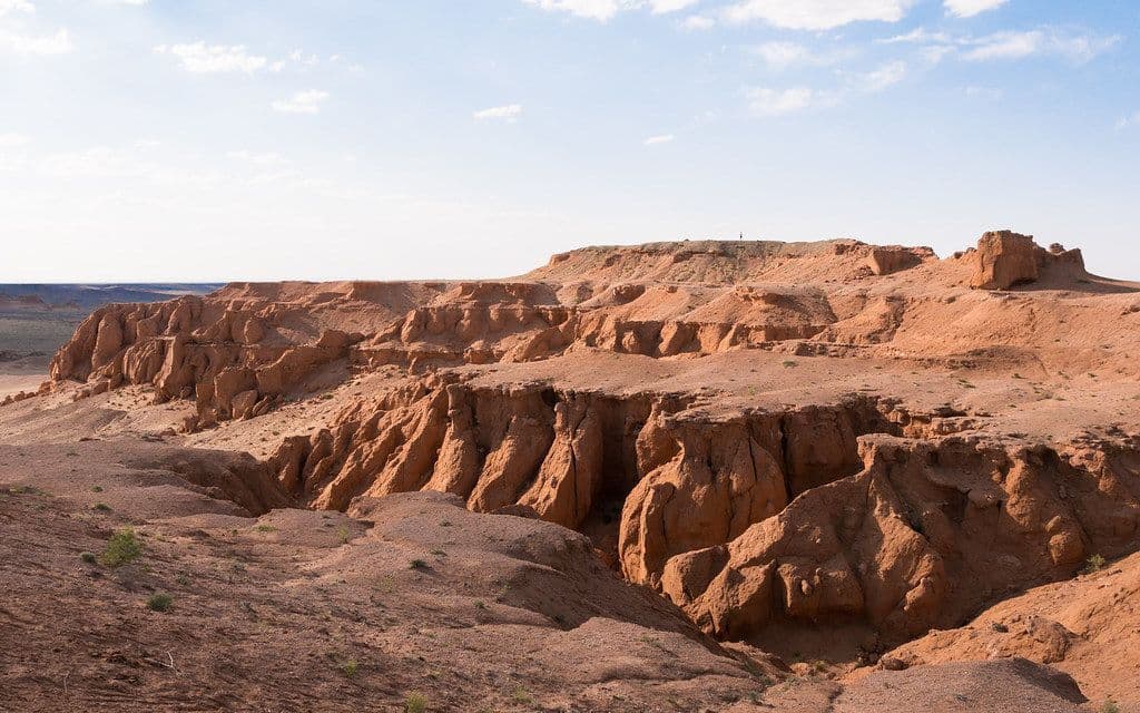 Una piccola sagoma si staglia in cima a una scogliera rosso-arancio, ammirando un vasto canyon eroso sotto un cielo azzurro pallido.