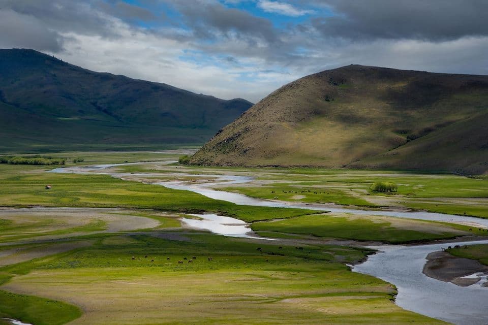 Un fiume serpeggia attraverso una valle lussureggiante e verde ai piedi di grandi colline ondulate sotto un cielo azzurro nuvoloso.