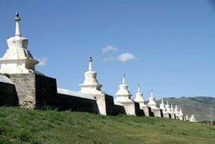 Una fila di stupa bianche si trova in cima a un lungo muro di pietra su un fianco di collina erboso sotto un cielo blu.