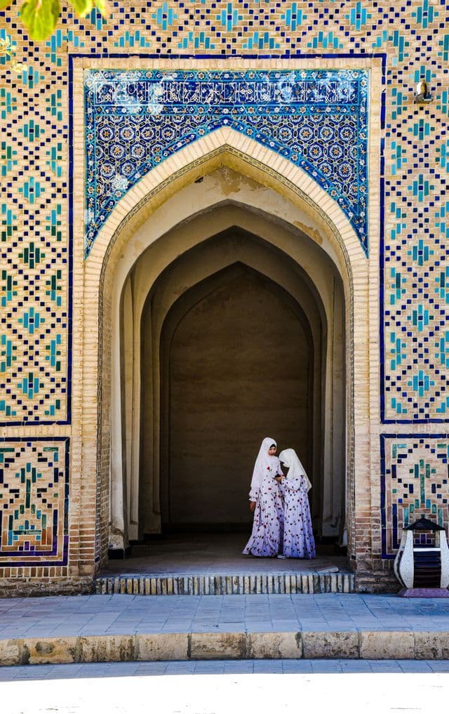 Zwei junge Mädchen in weißen Hijabs und passenden Kleidern stehen in einem kunstvollen Ziegelbogen, der mit blauen Mosaikfliesen verziert ist.