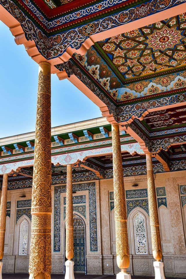 Des colonnes en bois finement sculptées soutiennent un portique avec un plafond coloré et à motifs sous un ciel bleu clair.