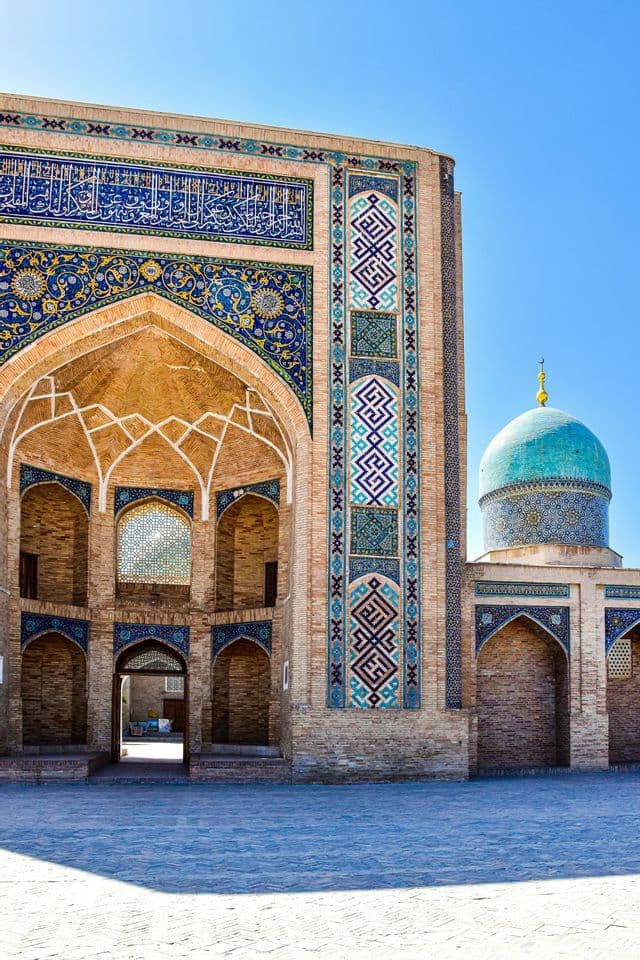 Ein kunstvolles Backsteingebäude mit filigranen blauen Mosaikarbeiten und einer türkisen Kuppel steht unter einem klaren blauen Himmel.