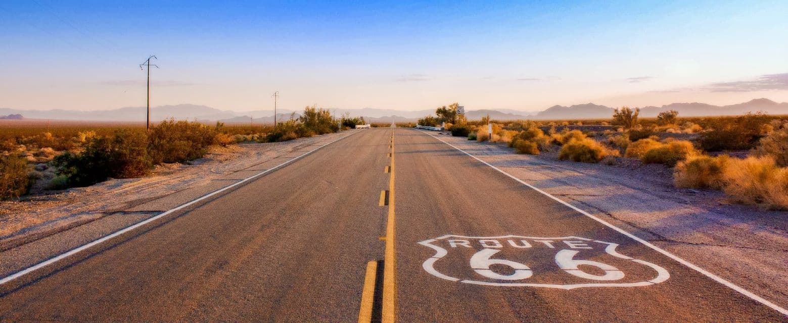 A Route 66 sign painted on the asphalt of an empty highway running through a desert landscape with mountains in the distance.