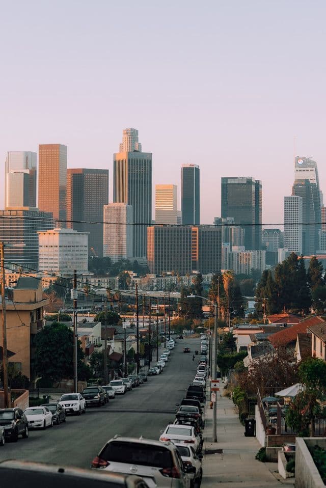 A view down a sloping residential street lined with parked cars, with a city skyline of skyscrapers in the background at sunset.