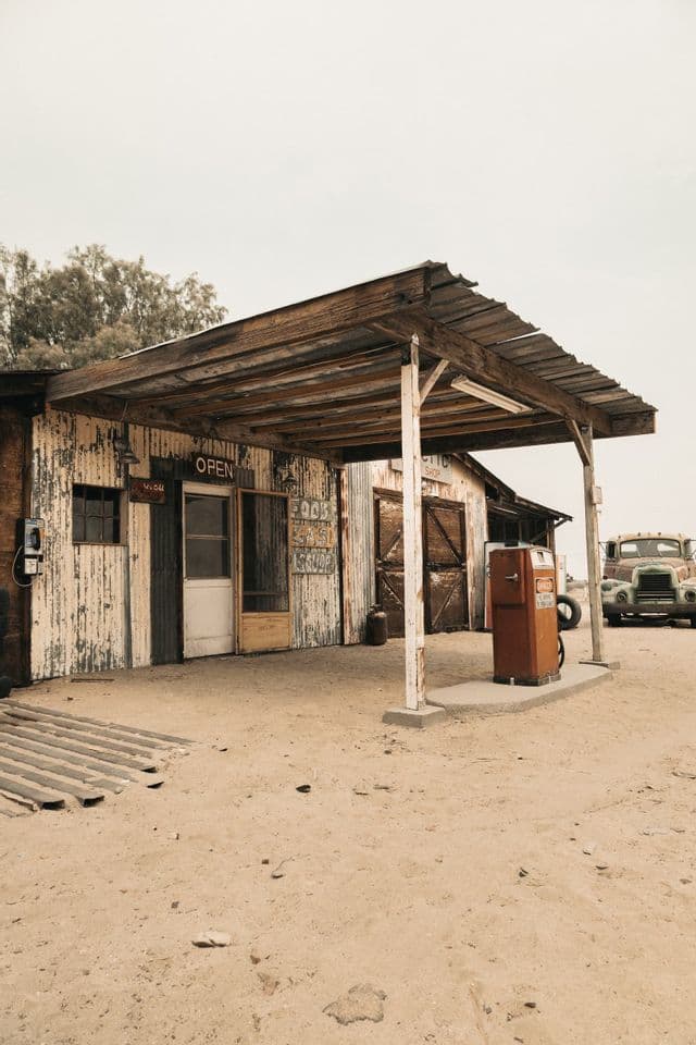 A rustic, weathered gas station with a vintage orange pump and an old truck parked on sandy ground.