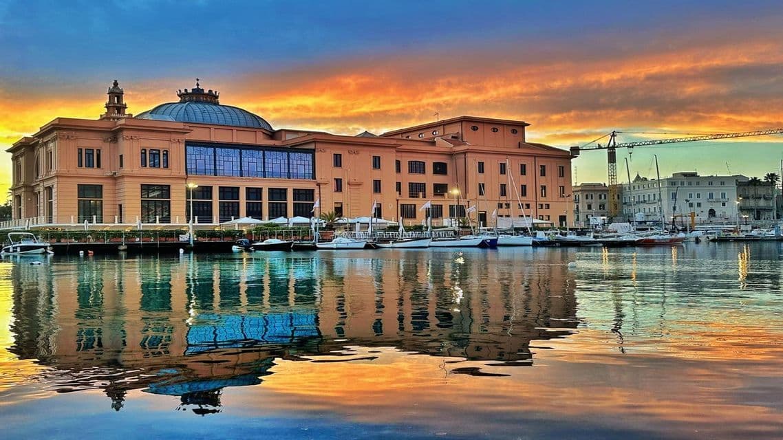 Un edificio ornato con una cupola di vetro che si riflette in un porto pieno di barche sotto un vivace cielo al tramonto.