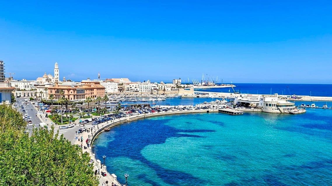 Una vista dall'alto di una città costiera con un porto turistico pieno di barche, una strada lungomare e acqua cristallina turchese sotto un cielo azzurro.