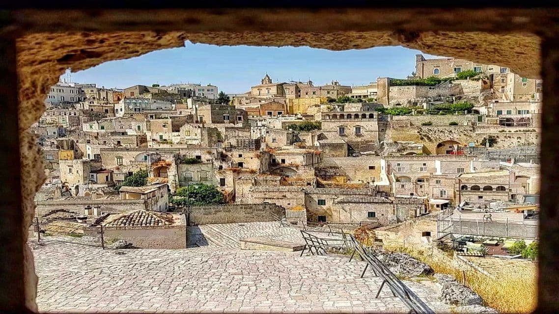 Una vista di una città storica con edifici in pietra su una collina, incorniciata da un arco in pietra grezza sotto un cielo blu.