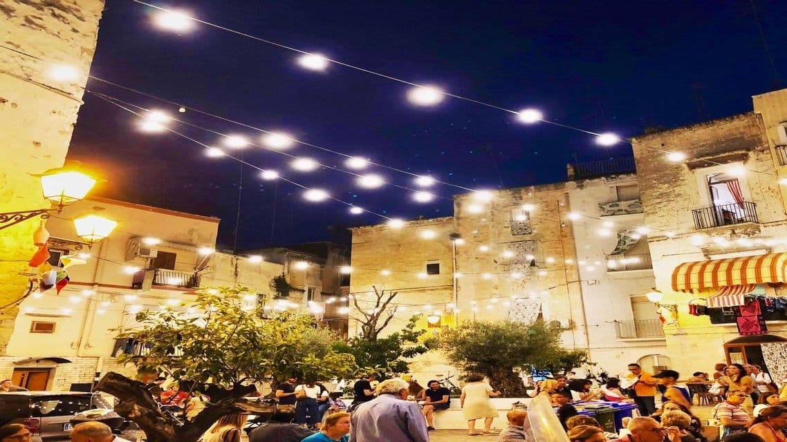 Una folla di persone si è radunata in una piazza di notte, illuminata da file di luci appese tra vecchi edifici.