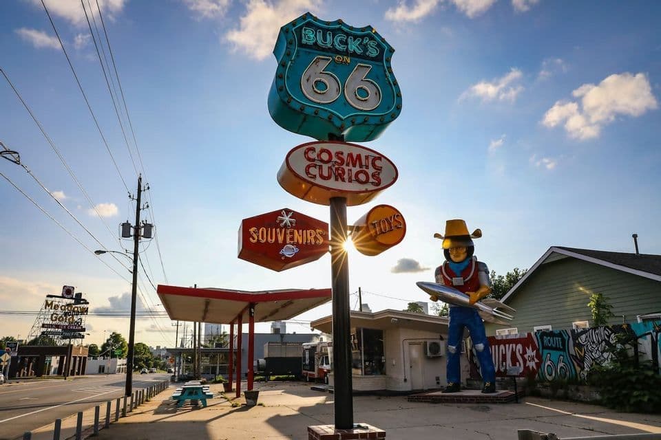 Vintage Route 66 signs and a large spaceman statue stand by a roadside shop under a sunny sky.