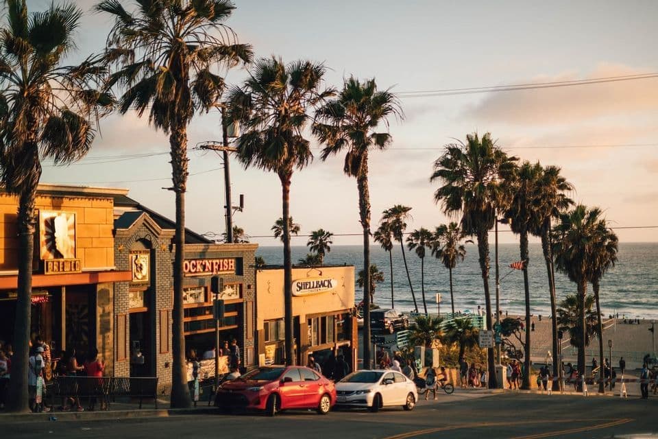 Una strada costeggiata da alte palme e negozi digrada verso una spiaggia sabbiosa e l'oceano al tramonto.