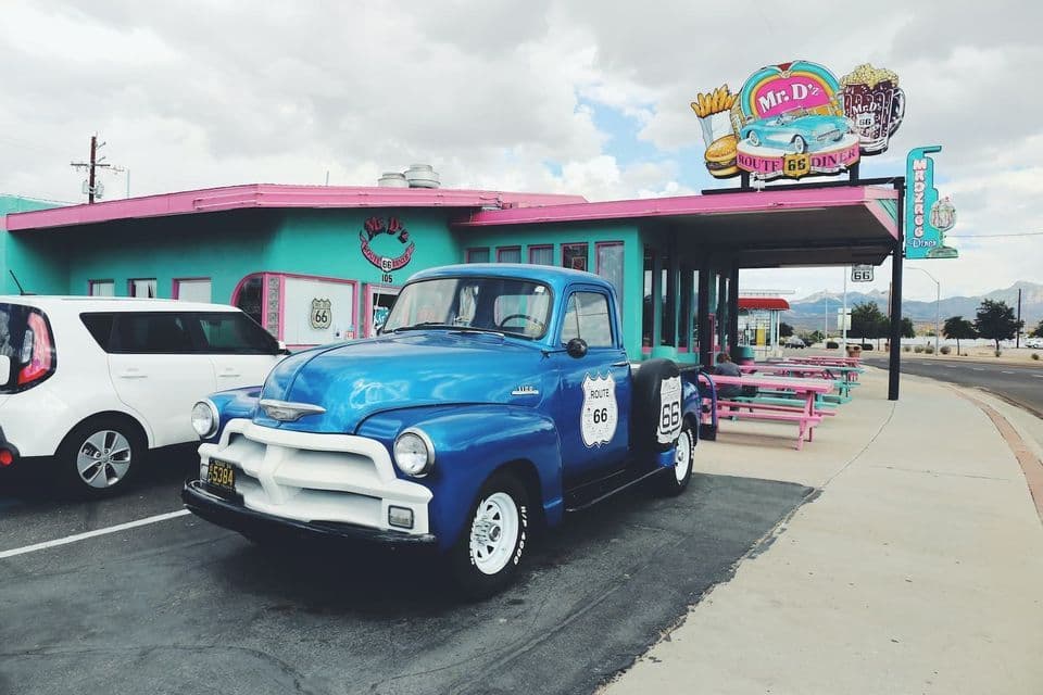A vintage blue pickup truck with a Route 66 logo is parked in front of a colorful, retro American diner on a cloudy day.