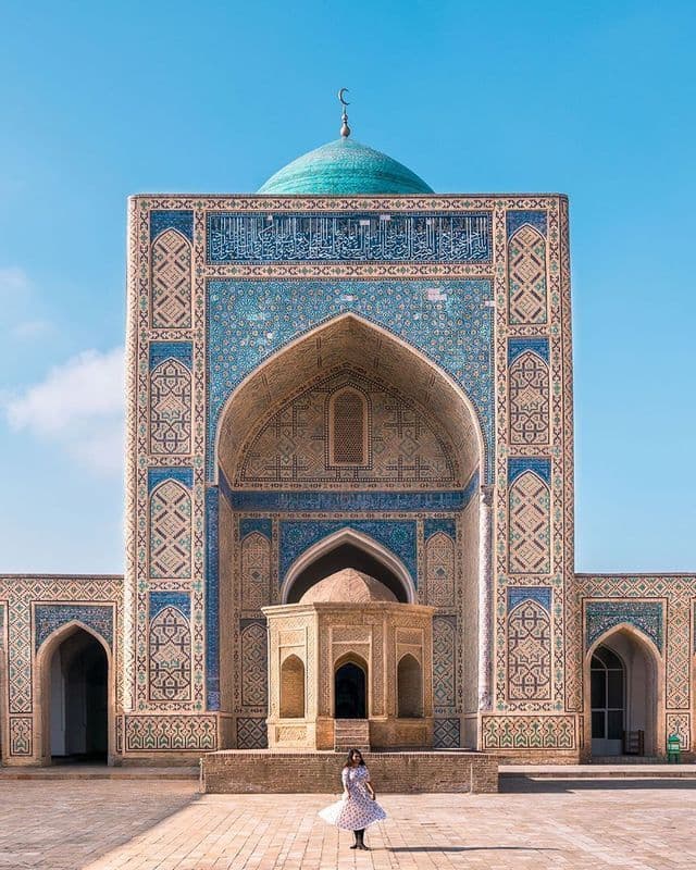 Una mujer con un vestido se encuentra en el patio de un edificio ornamentado con una cúpula turquesa y elaborados azulejos azules.