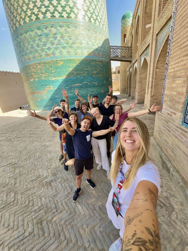 Eine WeRoad-Gruppenreise posiert für ein Gruppenselfie auf einer kopfsteingepflasterten Straße neben einem großen, türkisfarben gekachelten Turm.