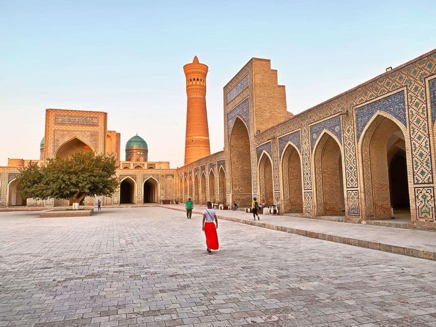 Une personne en jupe rouge traverse une grande cour en pierre vers des bâtiments en briques ornées avec un grand minaret.