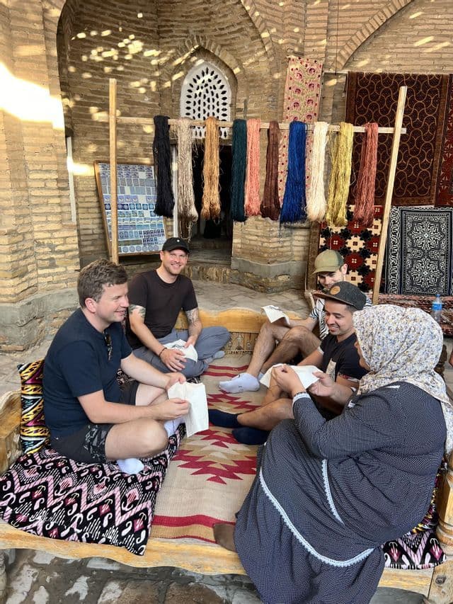 Un groupe WeRoad s'assied auprès d'une femme locale sur un banc, découvrant un savoir-faire artisanal dans un atelier aux fils colorés.
