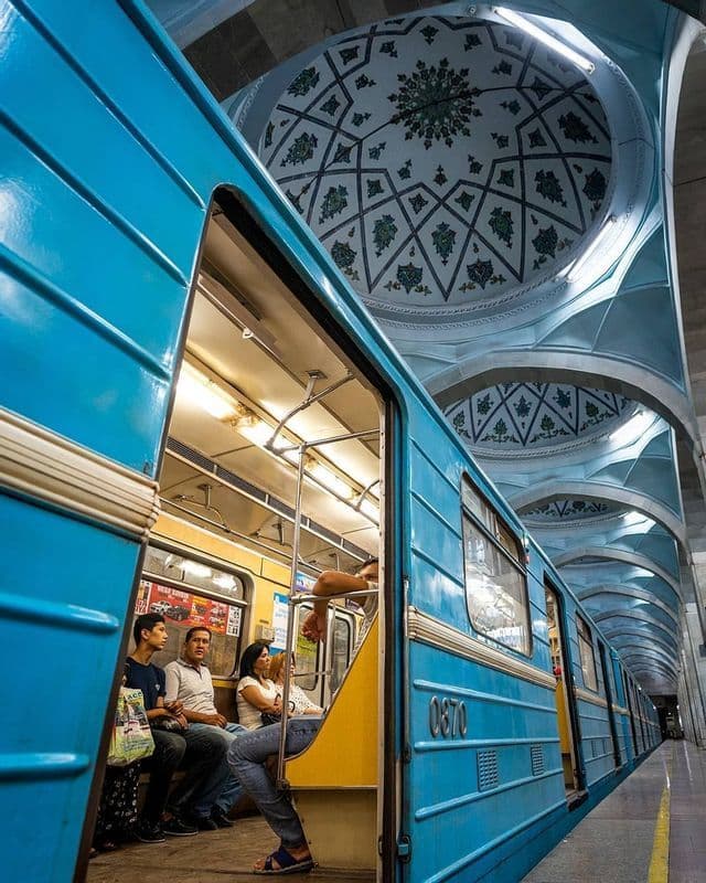 Un tren azul está detenido en el andén de una estación, revelando pasajeros en su interior, bajo un techo abovedado ornamentado con patrones geométricos.