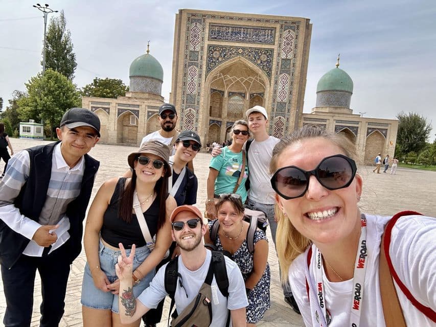 Eine WeRoad-Gruppenreise posiert für ein lächelndes Selfie auf einem Platz vor einem historischen Gebäude mit türkisfarbenen Kuppeln.