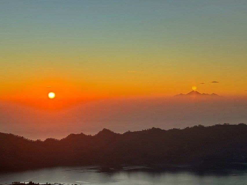 Un'alba vivace su una catena montuosa nebbiosa e un lago, con quello che sembra un secondo sole sorgere su una cima lontana.