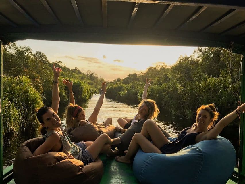 Un viaje en grupo de WeRoad de cuatro mujeres relajándose en pufs en un barco, navegando por un río selvático al atardecer.