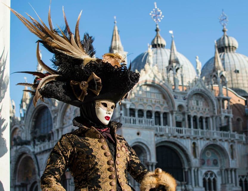 Una persona in un elaborato costume di carnevale veneziano con maschera e un grande cappello piumato si trova di fronte a una storica basilica a cupola.