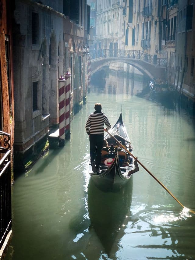 Un gondoliere con una maglietta a righe manovra una gondola con passeggeri attraverso uno stretto canale tra vecchi edifici.