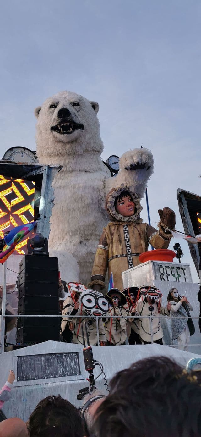 Un grande carro di carnevale con un gigantesco pupazzo di orso polare, una figura in parka e diverse persone che indossano maschere tradizionali.
