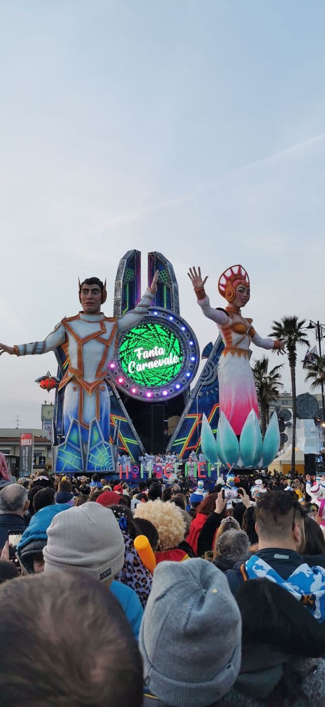 Una grande folla osserva un carro allegorico di carnevale con due figure futuristiche giganti sotto un cielo coperto.