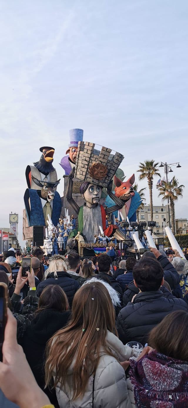 Una grande folla di persone assiste a un elaborato carro di carnevale con gigantesche figure di marionette durante una sfilata su una strada cittadina.