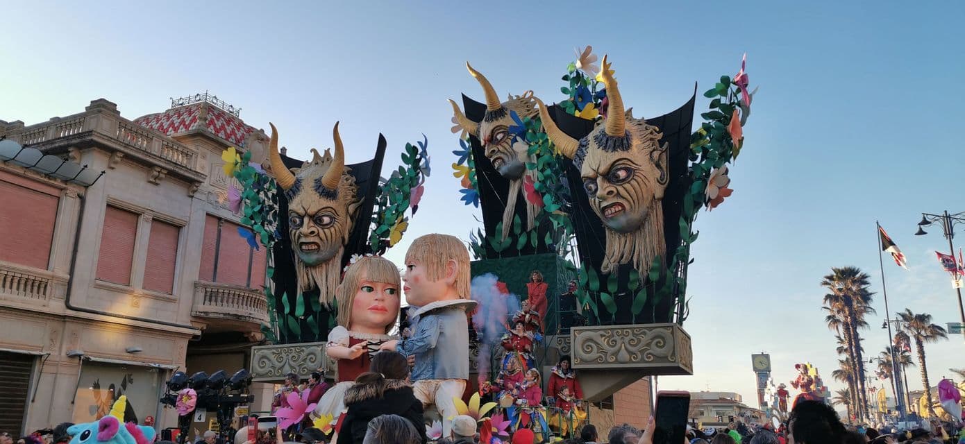 Un grande carro di carnevale con figure giganti in cartapesta di due bambini e tre demoni cornuti si muove lungo una strada affollata.