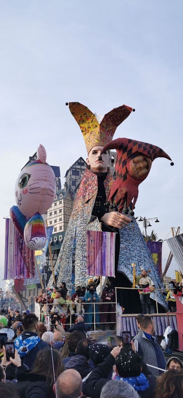Un enorme e colorato carro allegorico raffigurante un giullare domina una folla di spettatori durante una parata di strada.
