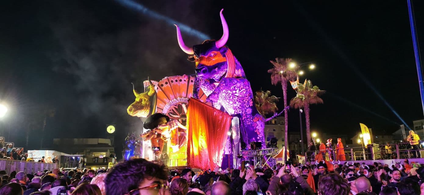 Un grande carro di carnevale illuminato a forma di toro viola sfila per una strada affollata di notte.