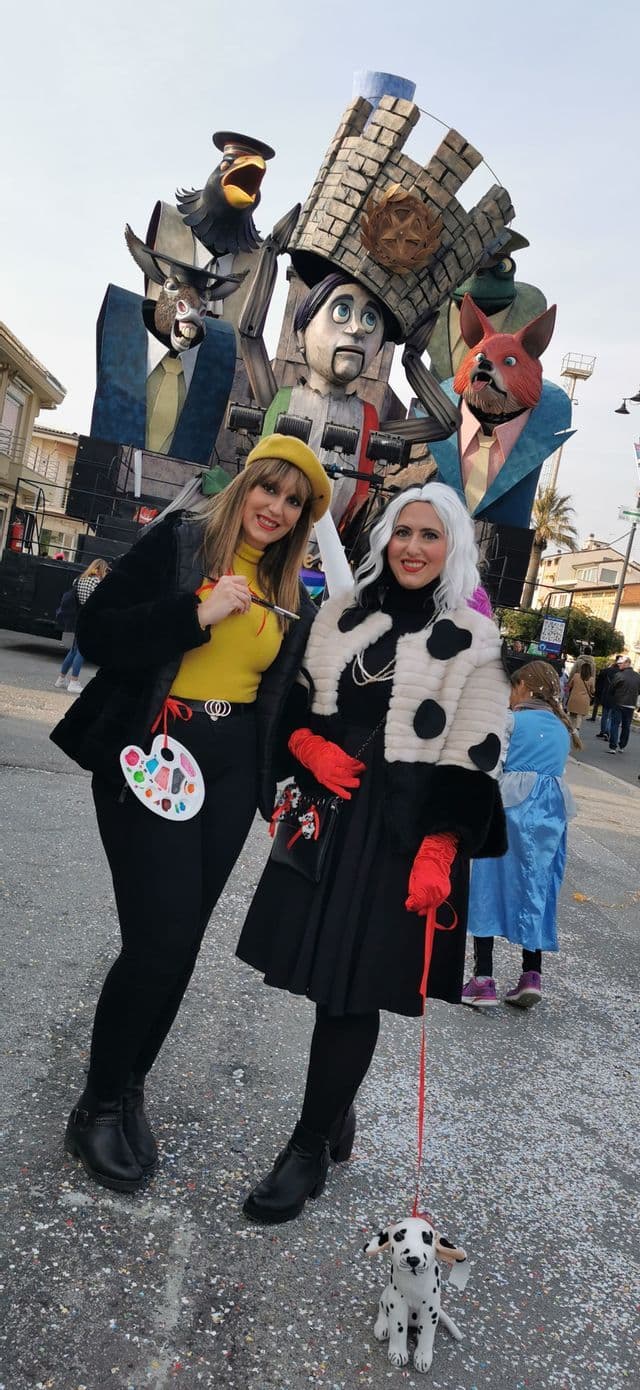 Due donne, una vestita da artista e l'altra da Crudelia De Mon, posano a un carnevale davanti a un grande carro allegorico.