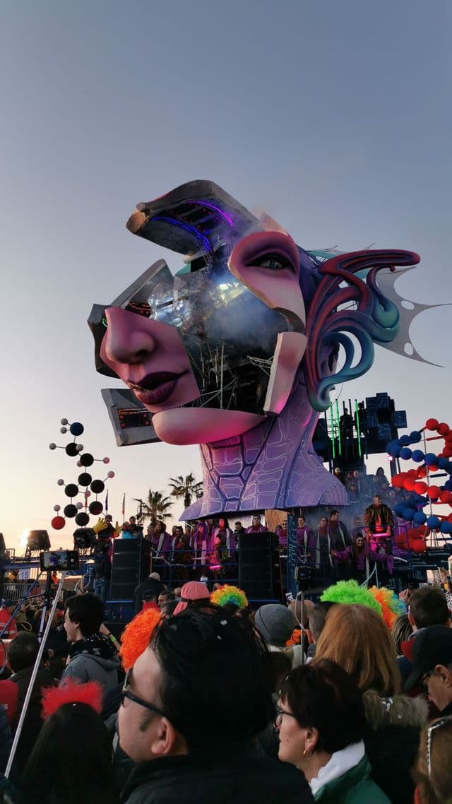 Un grande e elaborato carro allegorico con una testa di donna frammentata si staglia tra la folla a un carnevale.