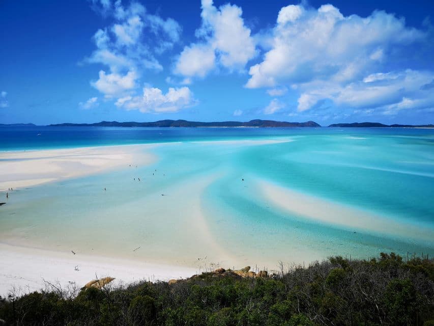 Vue plongeante d'une plage tropicale, avec des baigneurs dans une eau turquoise où le sable blanc tourbillonne sous un ciel bleu.
