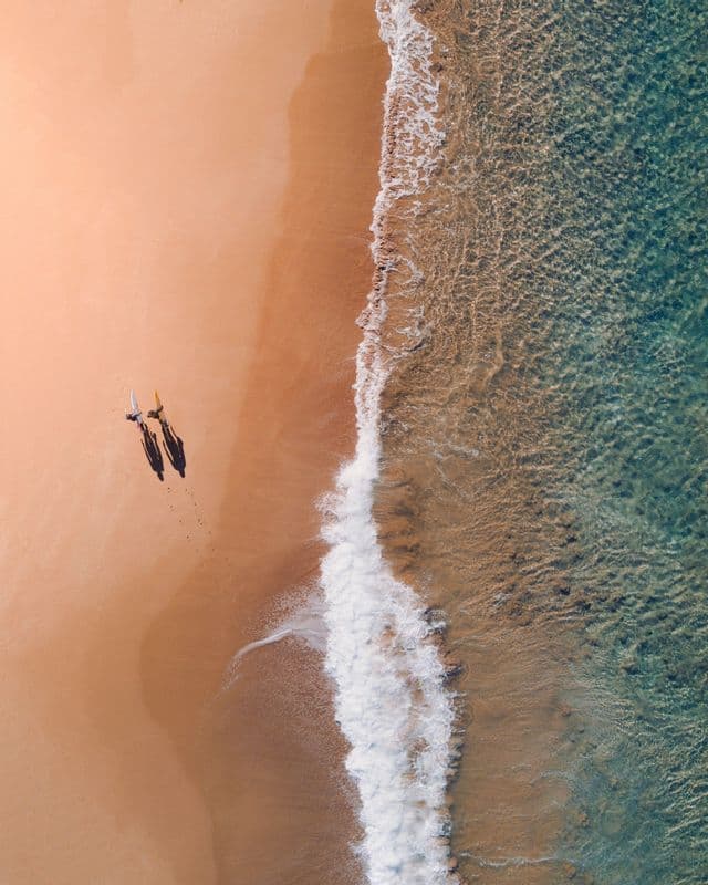 Vue aérienne de deux personnes avec des planches de surf marchant sur une plage de sable près des vagues de l'océan.