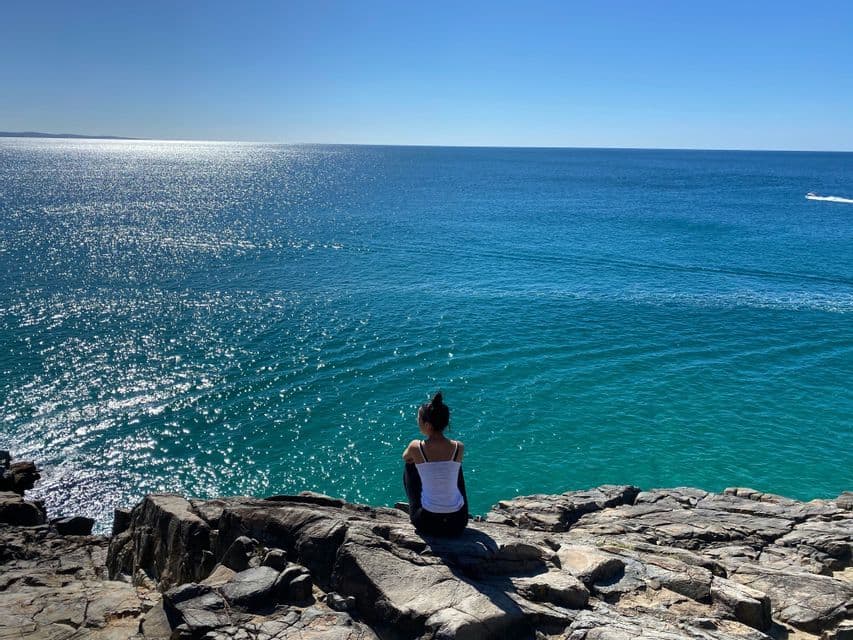 Une femme assise sur une falaise rocheuse, contemplant un vaste océan bleu chatoyant sous un ciel dégagé.