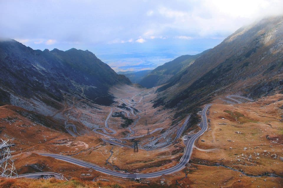 Veduta aerea di una strada tortuosa con numerosi tornanti che scende attraverso una vasta valle montana sotto un cielo nuvoloso.