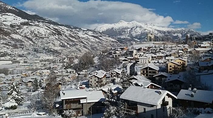 Un villaggio innevato si trova in una valle di montagna con imponenti cime innevate sullo sfondo sotto un cielo azzurro.