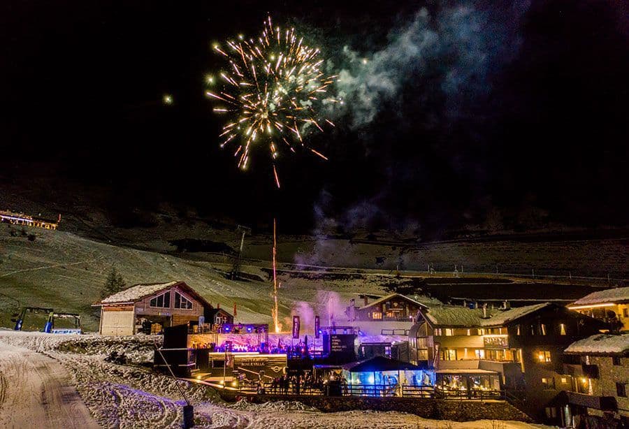 Fuochi d'artificio esplodono su un viaggio di gruppo WeRoad che festeggia di notte in una località montana innevata con chalet illuminati.
