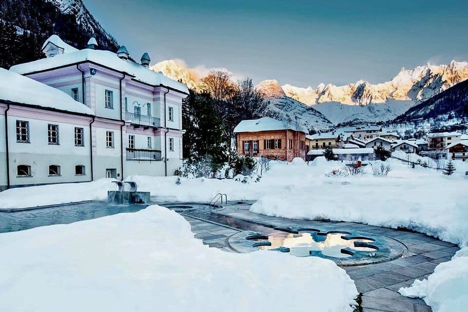Una piscina termale esterna fumante in un resort innevato, con montagne illuminate dal sole che si ergono sullo sfondo.