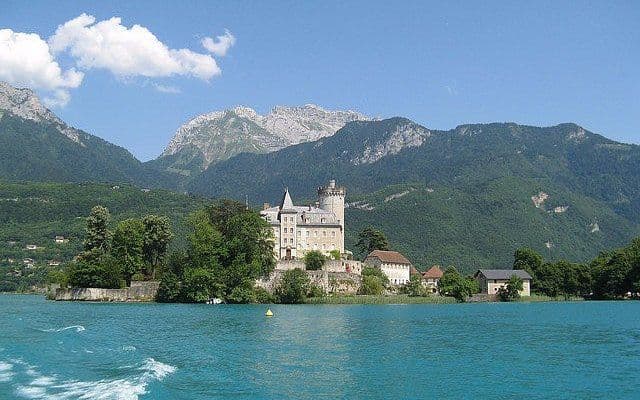 Un castello in stile medievale si trova su una piccola penisola in un lago turchese, con ripide montagne boscose sullo sfondo.