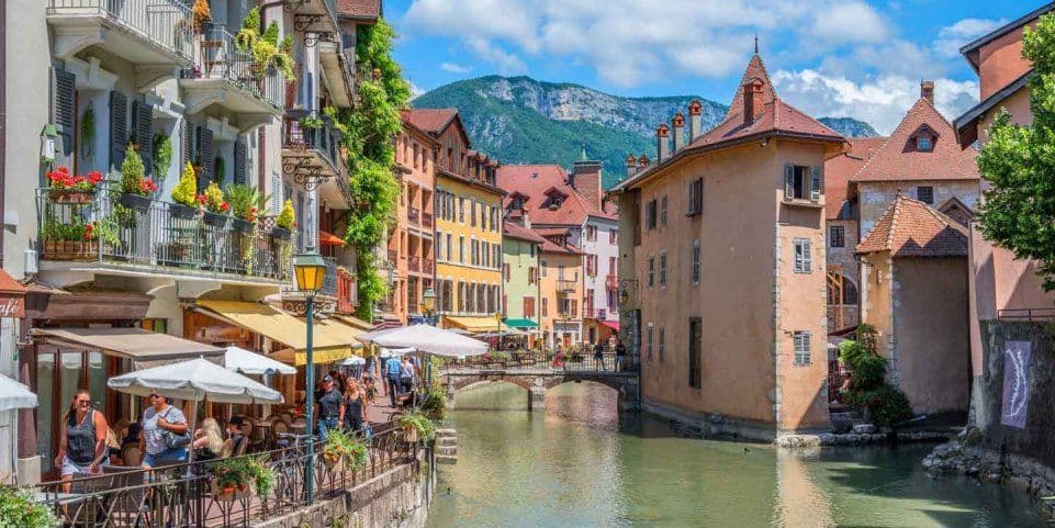 Colorati edifici storici con balconi fioriti costeggiano un canale in una città, con una grande montagna verde sullo sfondo.
