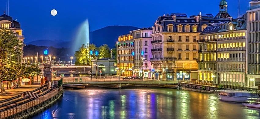 Edifici illuminati costeggiano un fiume di notte, con una grande fontana e una luna piena nel cielo blu profondo.
